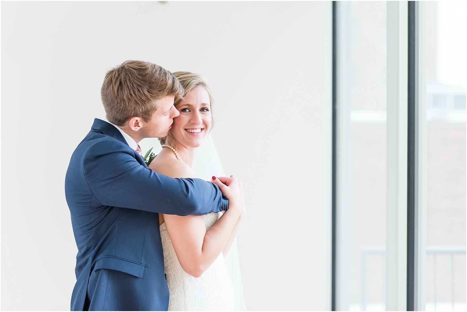 Groom wrapping his arms across his bride and she smiles at the glass box