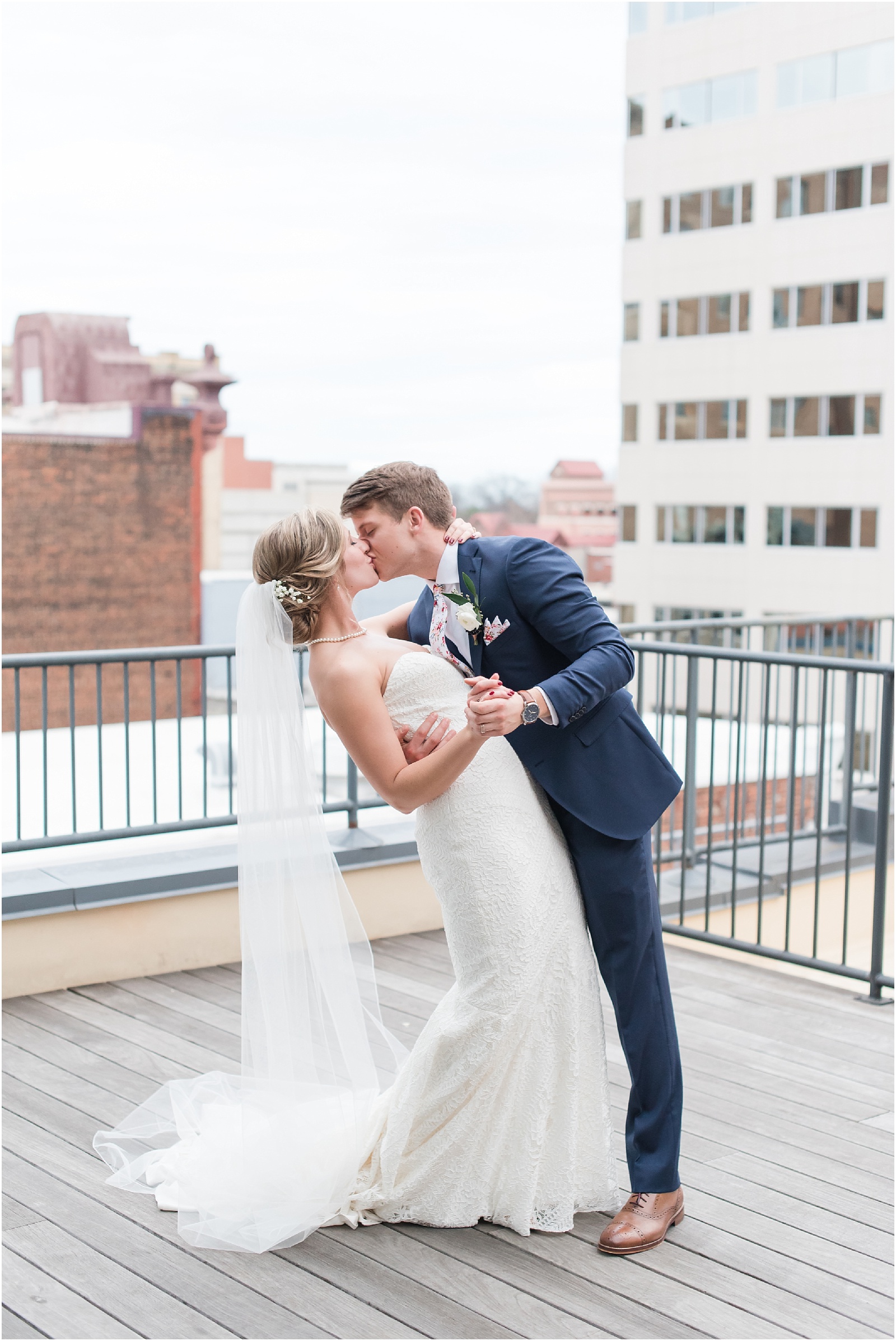Bride and groom in a dip kiss on rooftop patio with a view of downtown Raleigh at the glass box