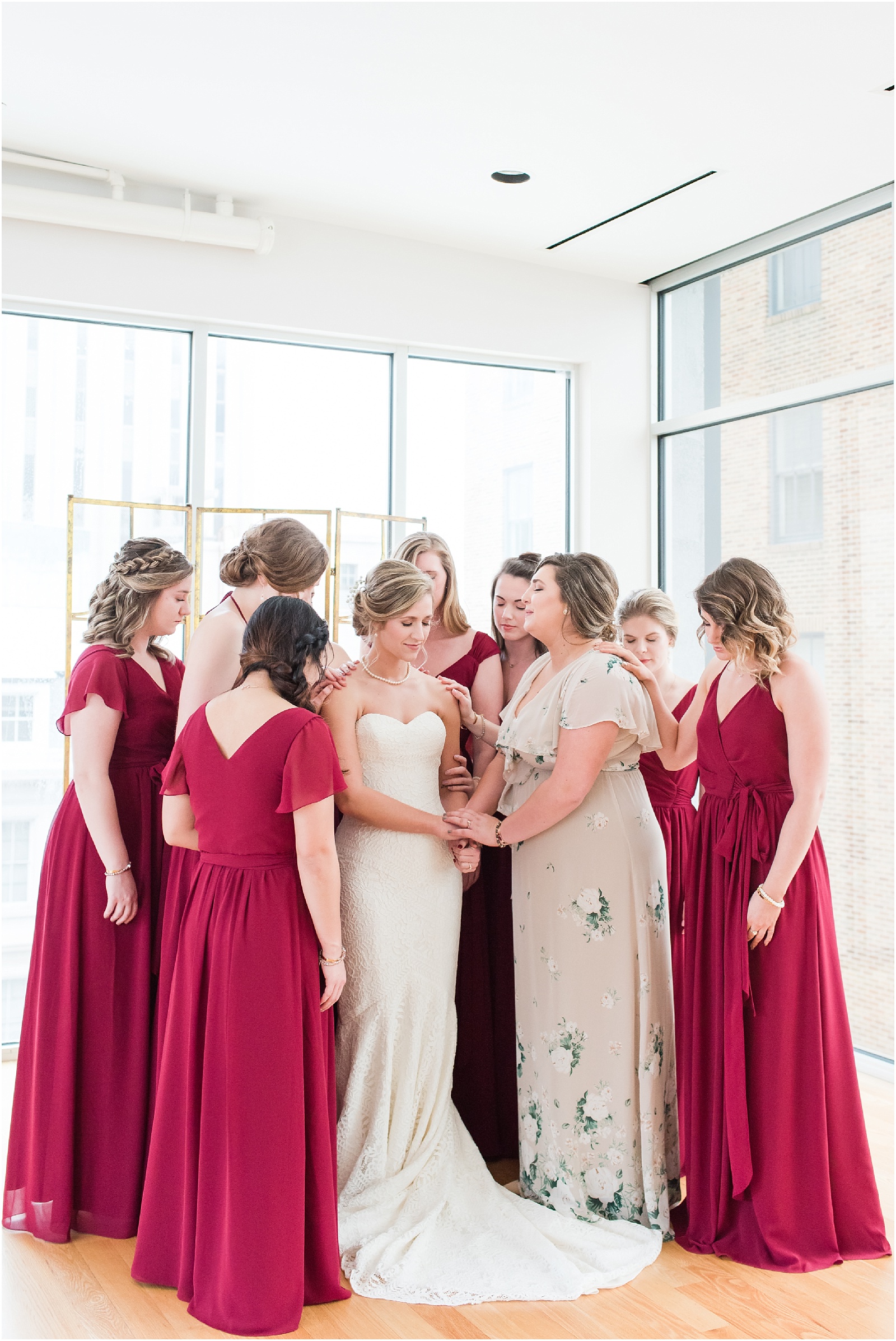 Bride in her bridal gown with bridesmaids in red full length dresses praying with bride at the glass box