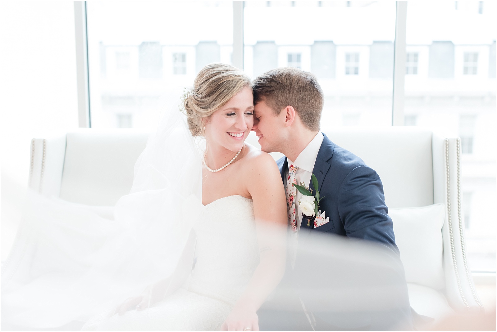 Bride and Groom sitting on white couch with long veil swooping across at the glass box