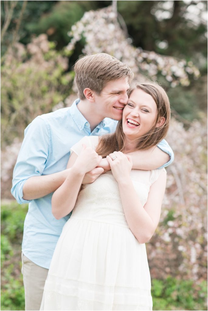a couple at an engagement session standing in front of a cherry blossom tree with the groom standing behind and holding his fiance wearing blue button up with the bride wearing a cream colored dress laughing