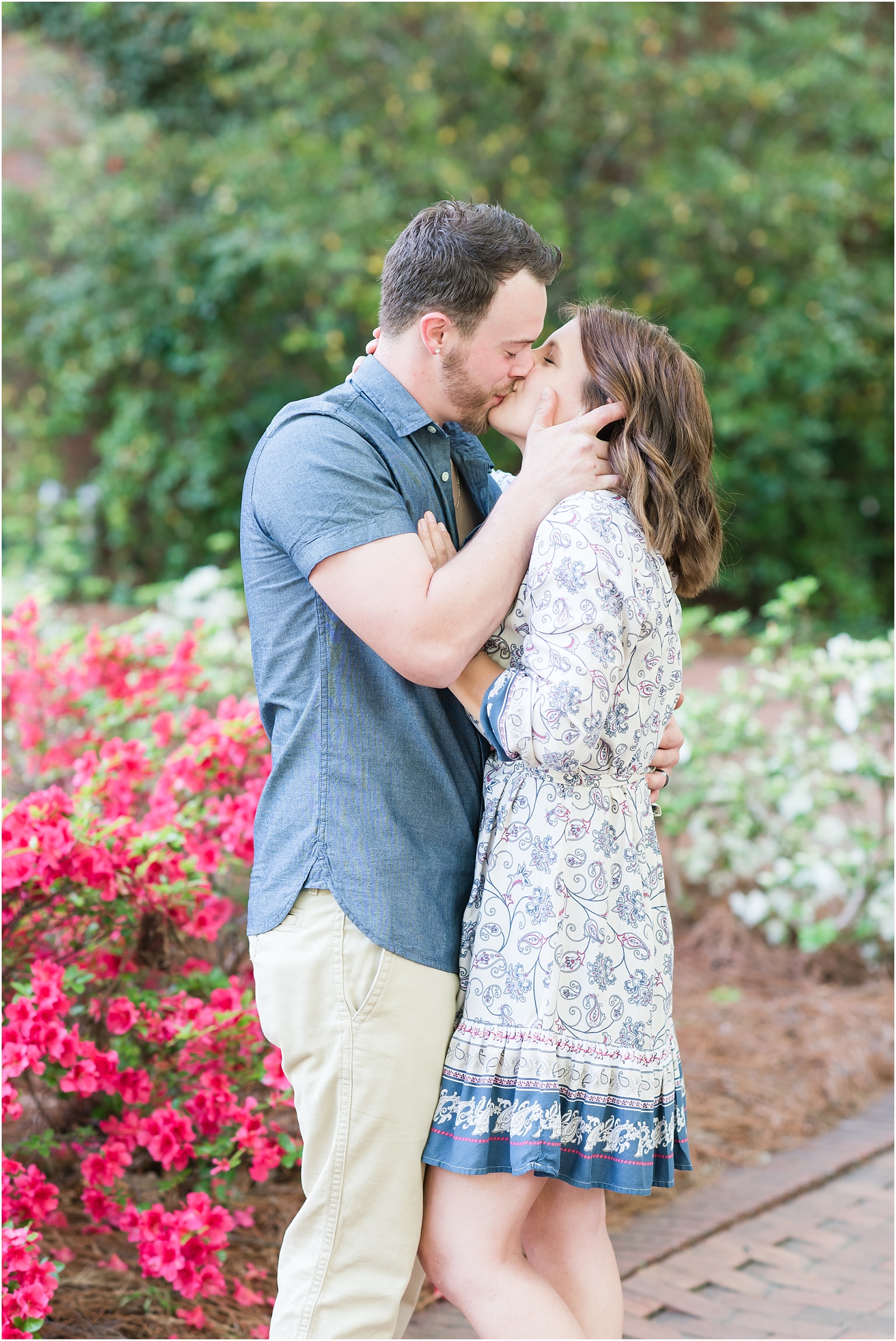 young couple kissing in front of flowering bushes and ivory wall at Elon College