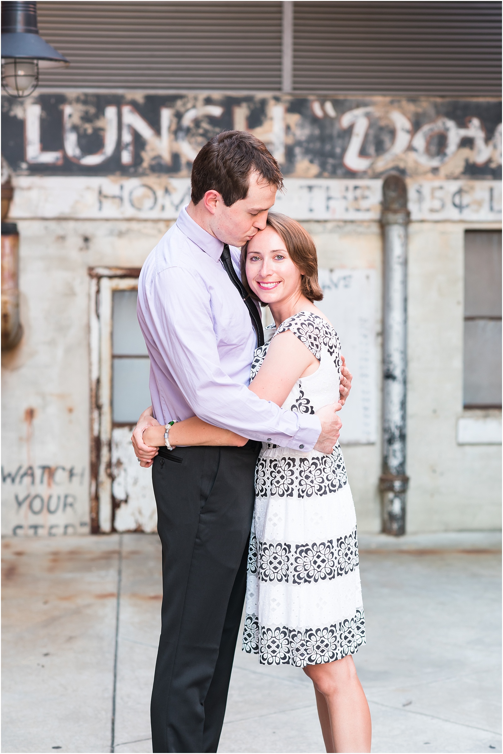 young man kissing woman on forehead in front of a rustic wall at the American Tobacco Campus