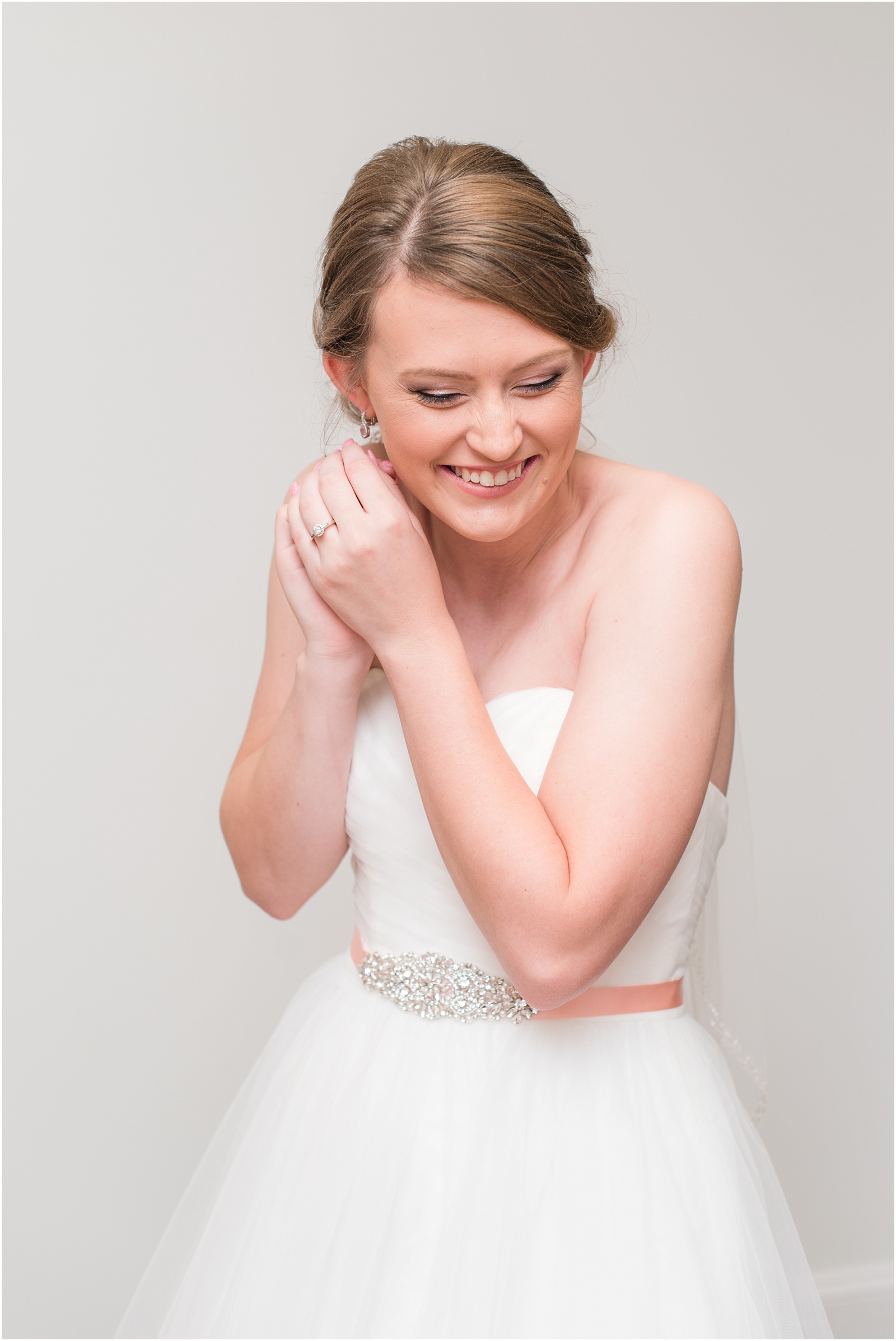 bride in white wedding dress with pink ribbon putting on earrings with a smile to her face grey details with touches of soft pink with rinks and earrings at City Lake Baptist Church