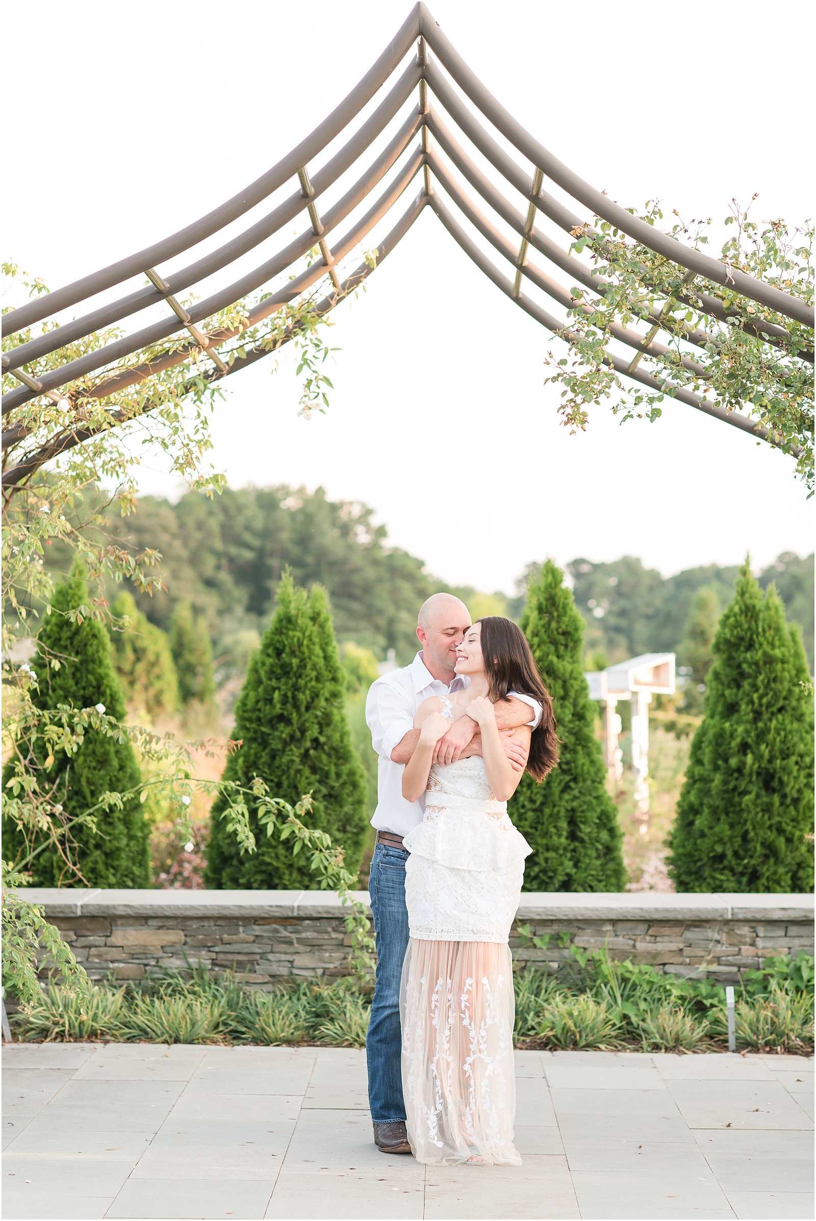 a man wearing a white snuggling up from behind with a woman wearing a white dress with dusty blue heels underneath a vine arbor at JC Raulston Arboretum