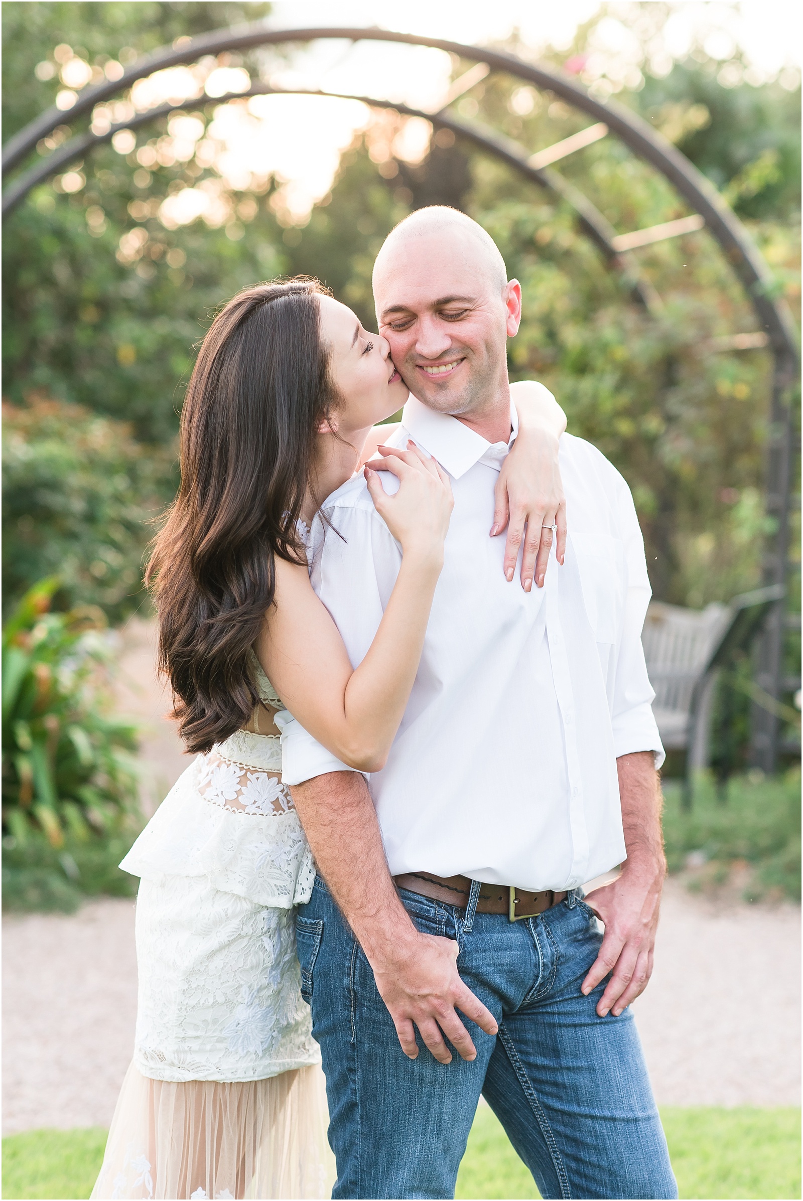 a women wearing a white dress hugging man wearing white shirt with dark blue jeans from behind kissing his check at JC Raulston Arboretum
