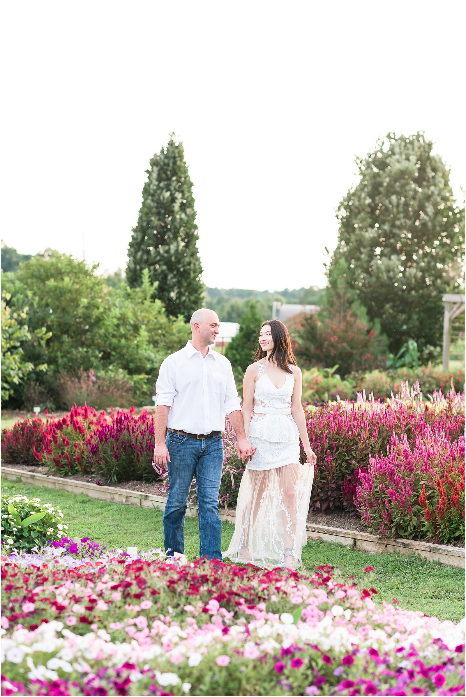 man wearing a white shirt and woman wearing a white dress walking hand in hand through a flower field at JC Raulston Arboretum