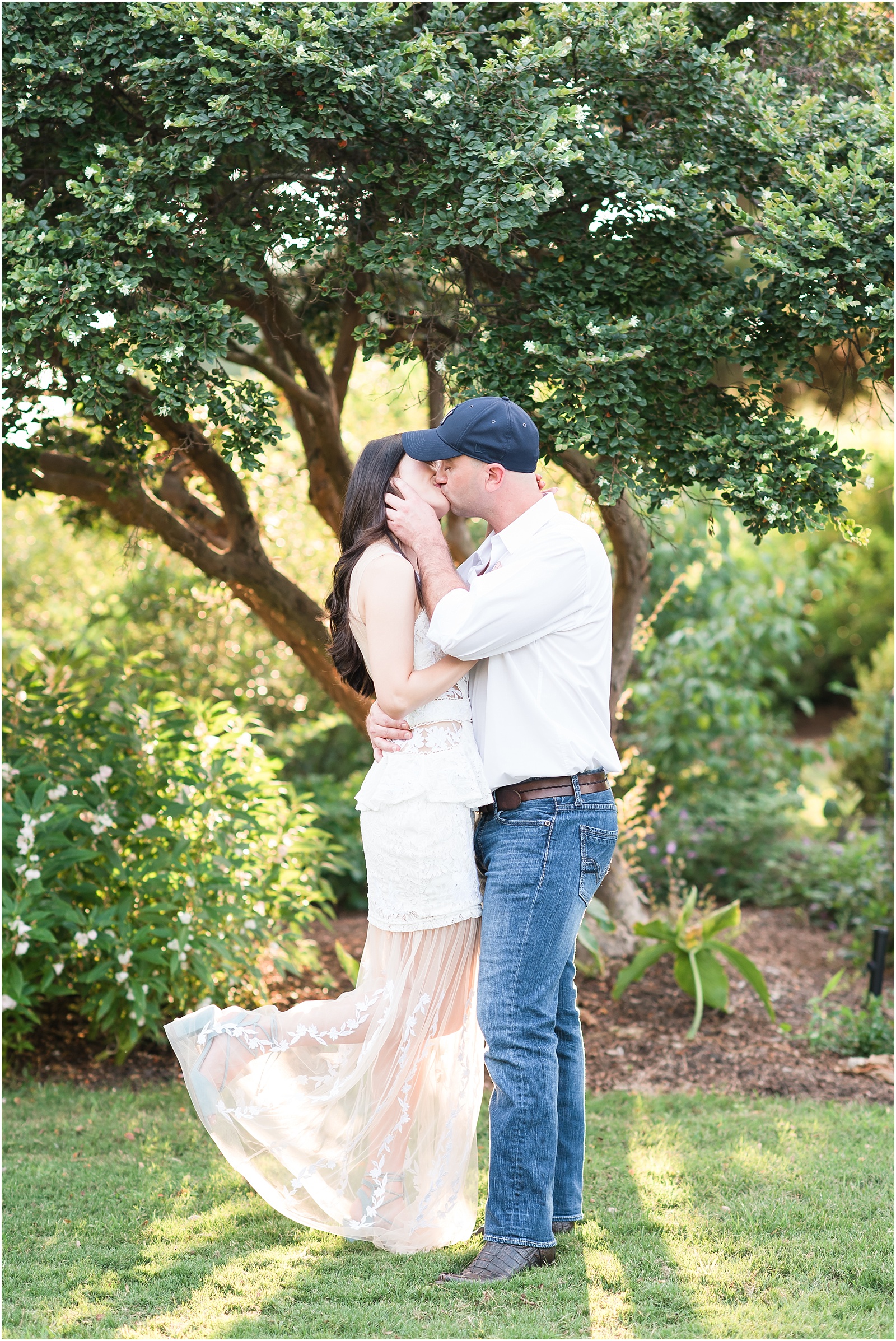 man wearing a white shirt and dark blue jeans cupping his fiance's check and giving her a kiss while she wearing a white dress and dusty blue heels pops her foot up at JC Raulston Arboretum
