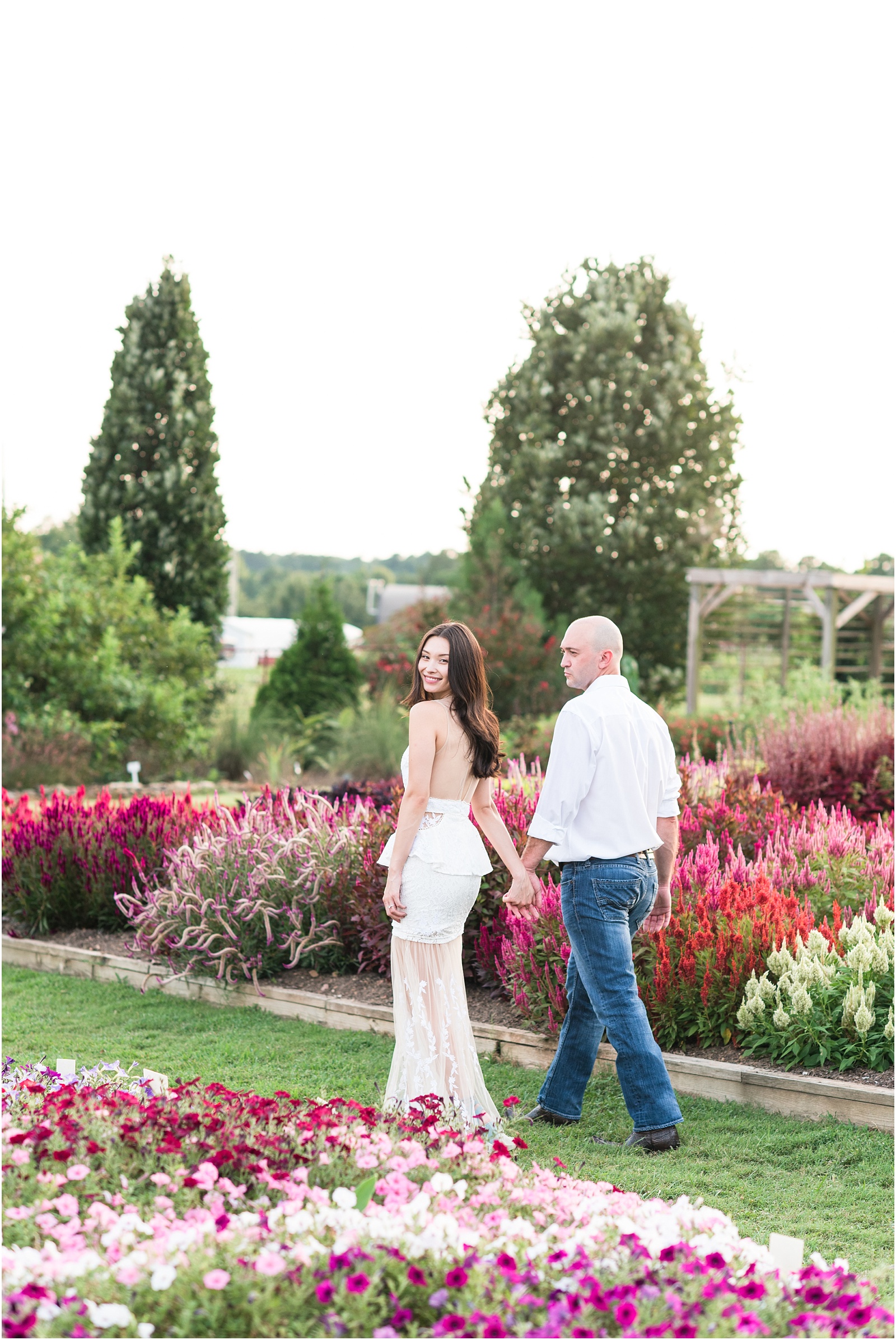 a man wearing blue jeans and a white shirt is walking hand in hand with his fiance who is looking back at the camera wearing a white dress with dusty blue heels through a flower field at JC Raulston Arboretum
