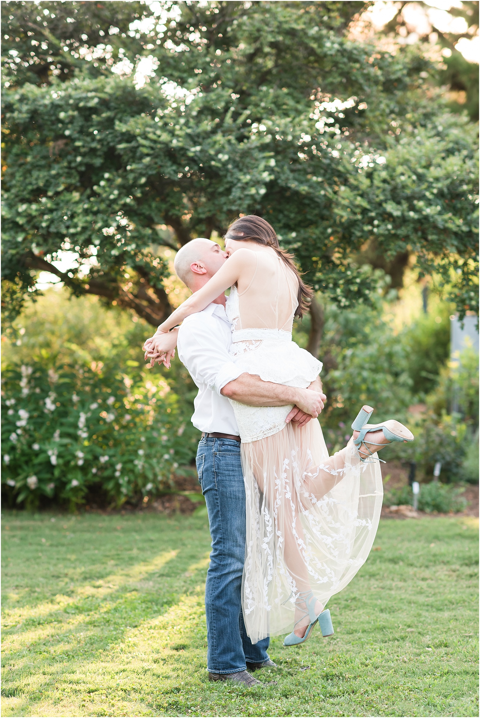 a man wearing a white shirt with dark blue jeans lifting up and kissing a woman wearing a white dress with dusty blue heels at JC Raulston Arboretum