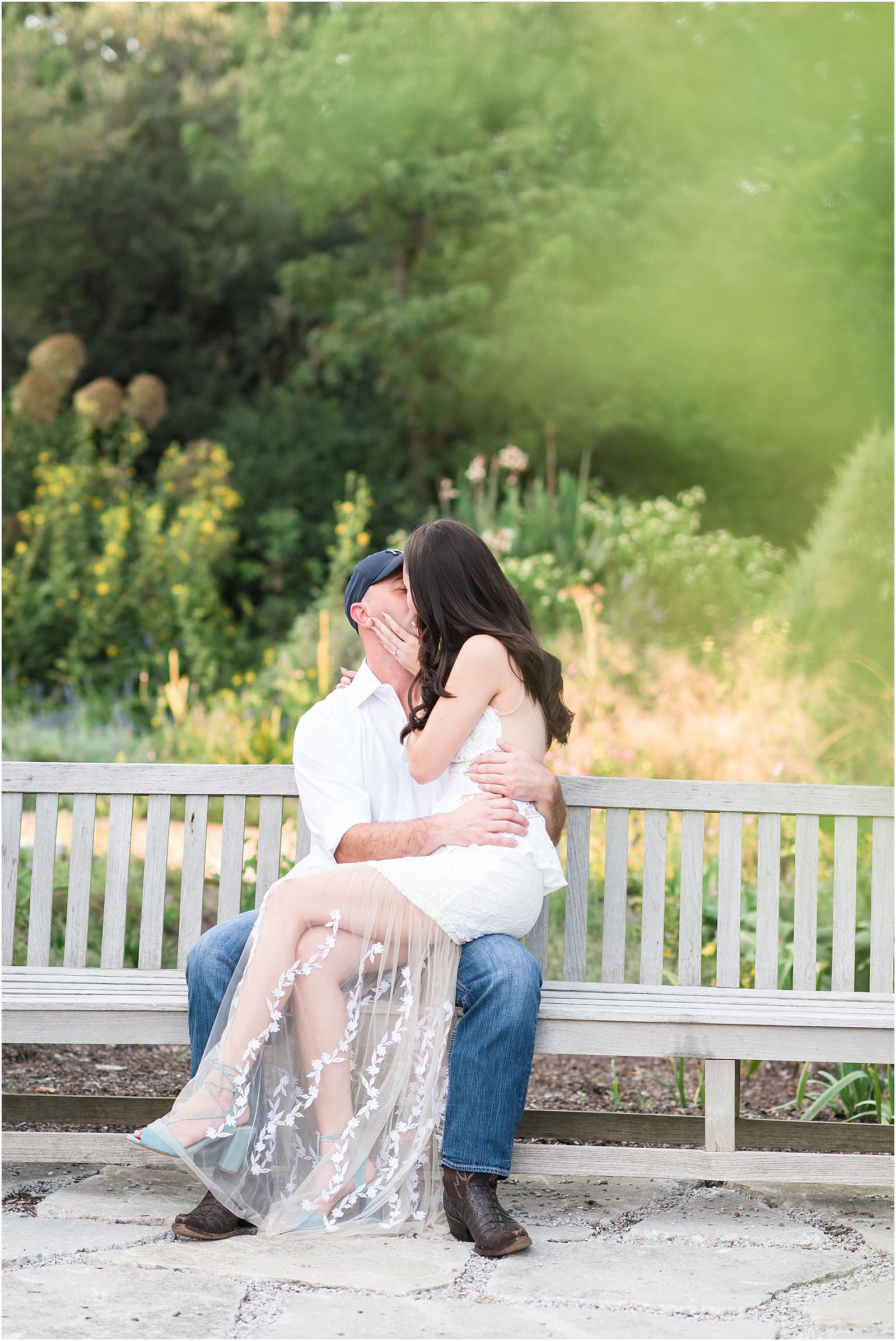 a woman wearing a white dress with dusty blue heels sitting on a man wearing a white shirt with dark blue jeans lap giving him a kiss at JC Raulston Arboretum