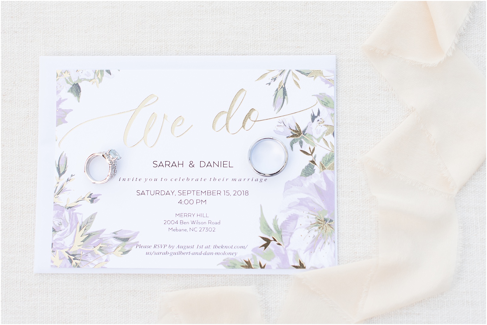 a purple, green, and gold wedding invitation to Merry Hill Weddings with beige ribbon with a blue stoned engagement ring and sliver wedding band on top