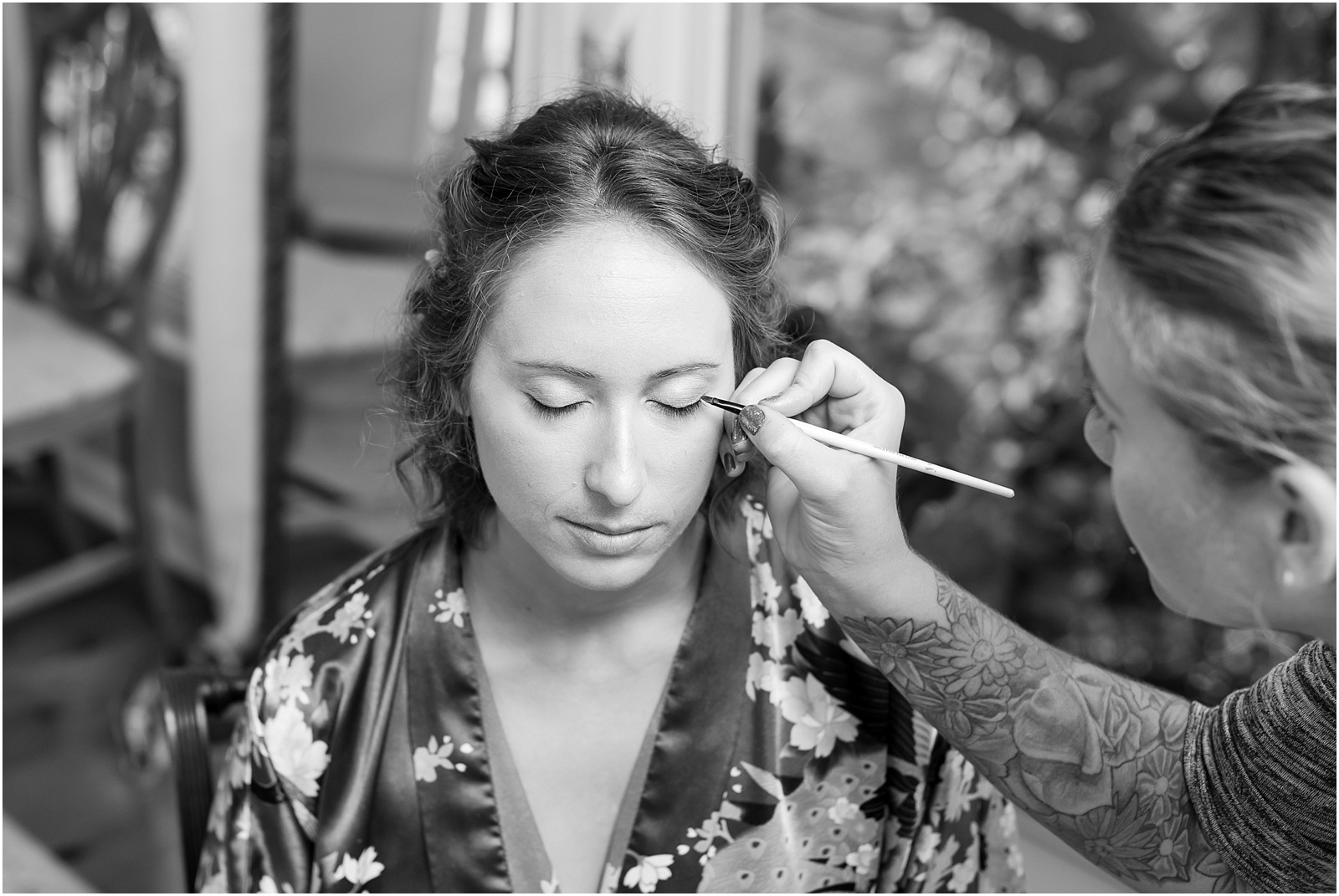 a black and white photojournalistic photograph of bride, wearing a bridal robe, getting her eyeliner done by a makeup artist