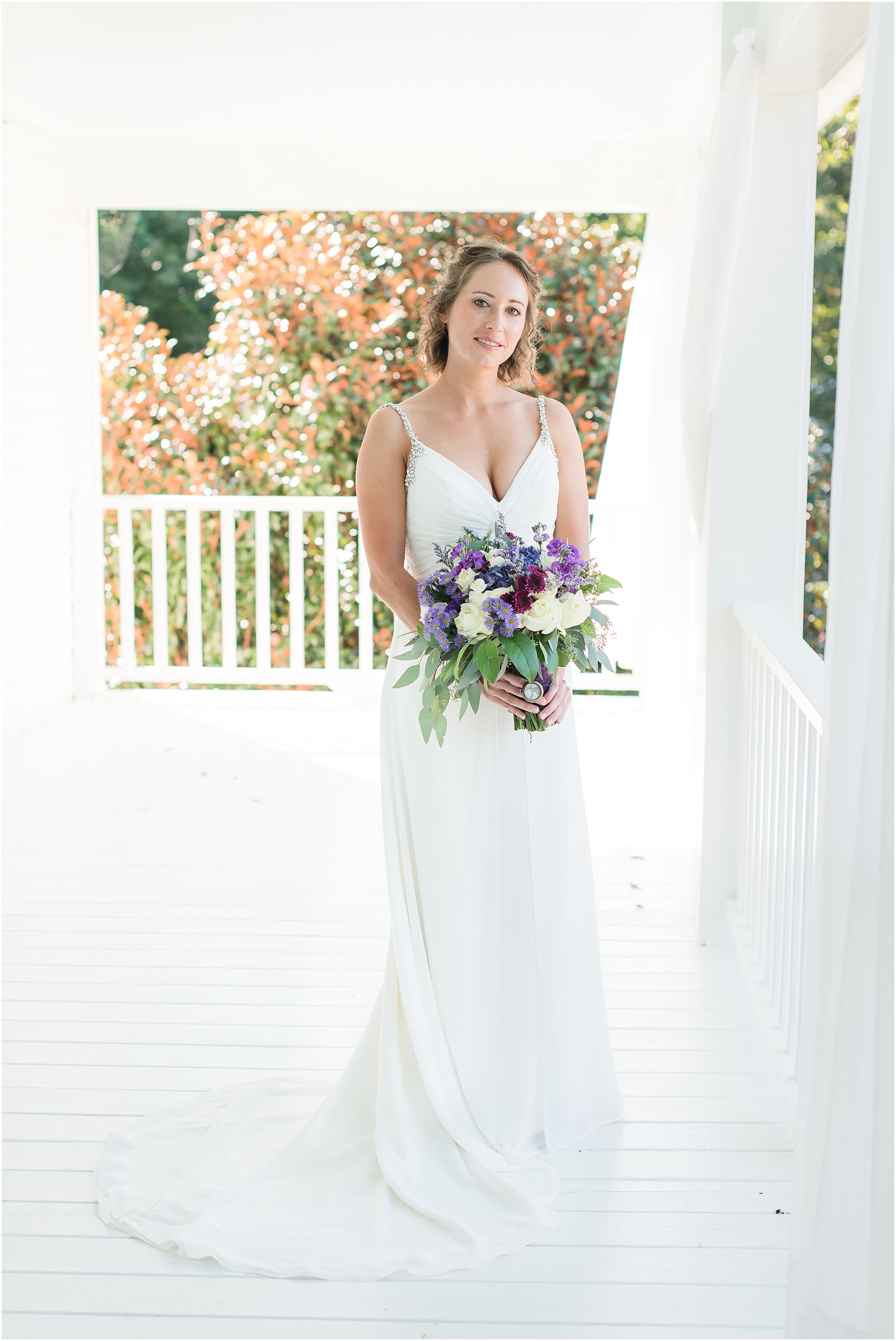 bride wearing white dress with sliver beaded straps holding a purple/pink/green wedding bouquet on the porch of Merry Hill