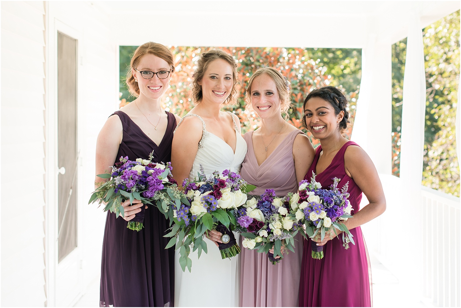 a photograph of a bridal and bridesmaids wedding bouquets filled with purple, pink, blue, green, and white flowers on the porch of Merry Hill Weddings