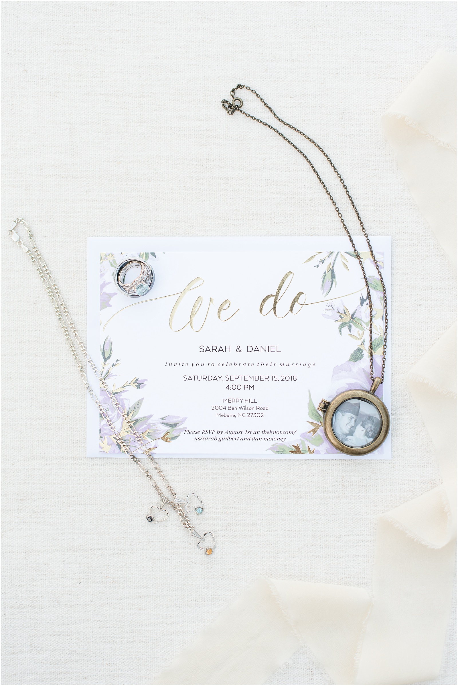 a purple and gold wedding invitation surrounded by a blue stoned engagement ring and sliver wedding band with a locket and sliver necklace