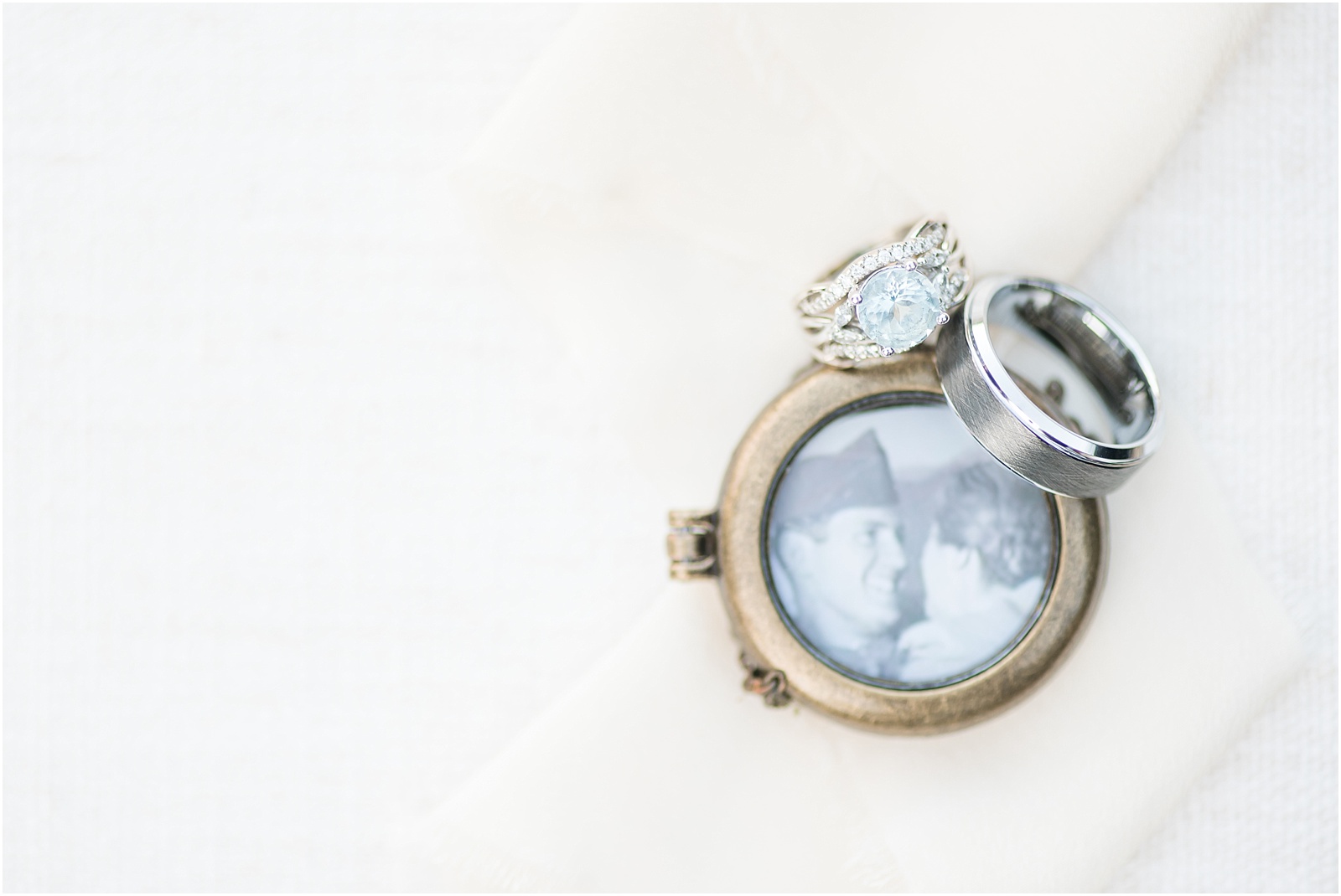 a blue stoned wedding ring sitting next to a sliver wedding band on top of a locket with an old photograph inside on top of a beige background