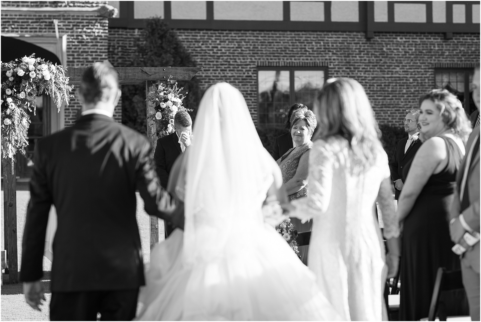 bride walking down the aisle with mom and dad with the focus on the groom holding a handkerchief up to his eyes crying at Sedgefield Country Club