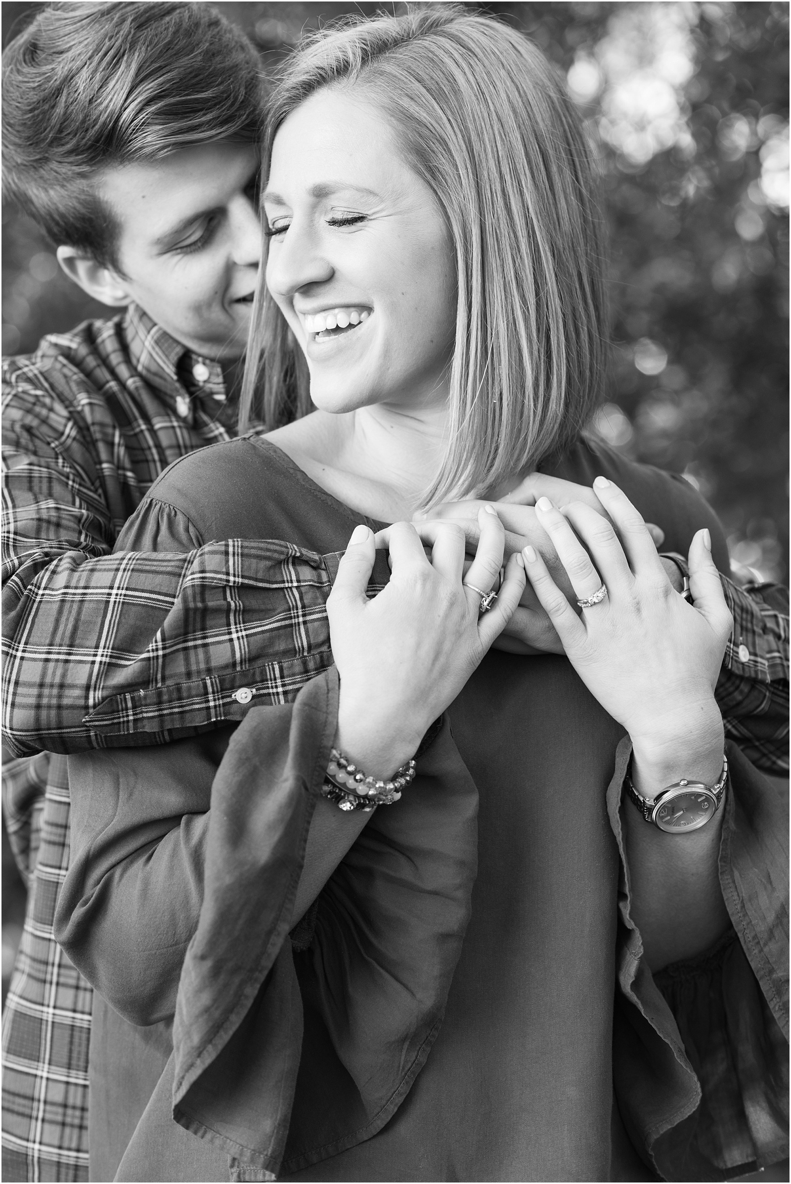a black and white photograph of a groom holding onto his bride from behind kissing her cheek and making her laugh for choosing your engagement session