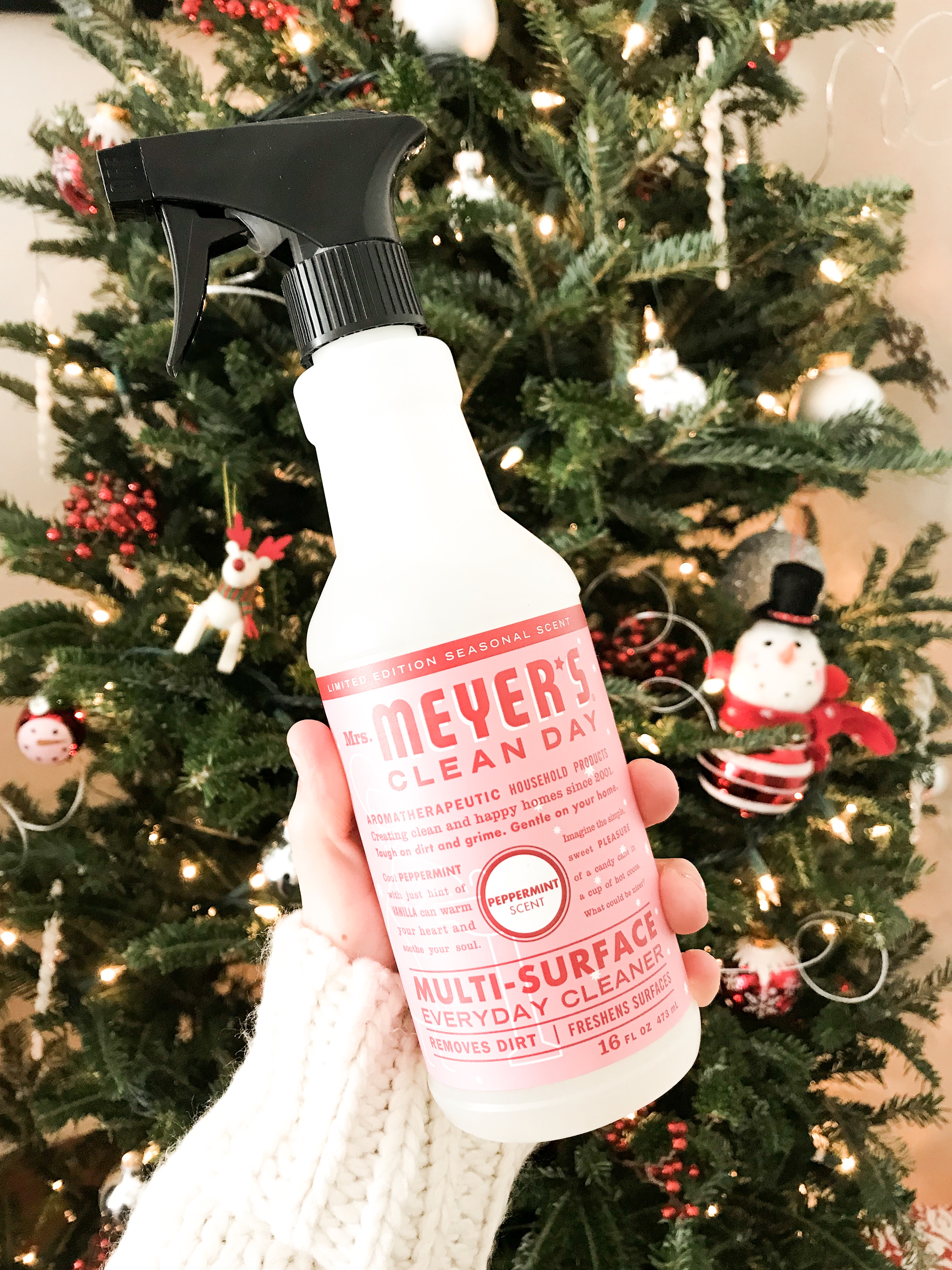 a woman holding a bottle of Mrs. Meyer's multi surface cleaner in front of a snowman christmas tree